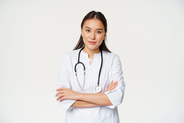 Retrato de mujer asiática médico, brazos cruzados, de pie en uniforme médico y estetoscopio, sonriendo a la cámara, fondo blanco.