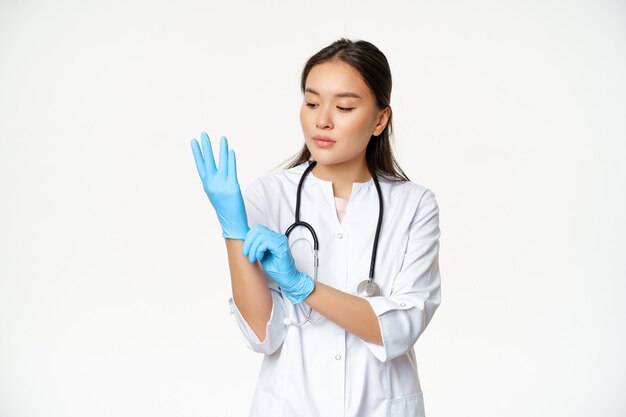 Retrato de mujer asiática médico se pone guantes de goma para examinar al paciente en la clínica, de pie con uniforme sanitario sobre fondo blanco.