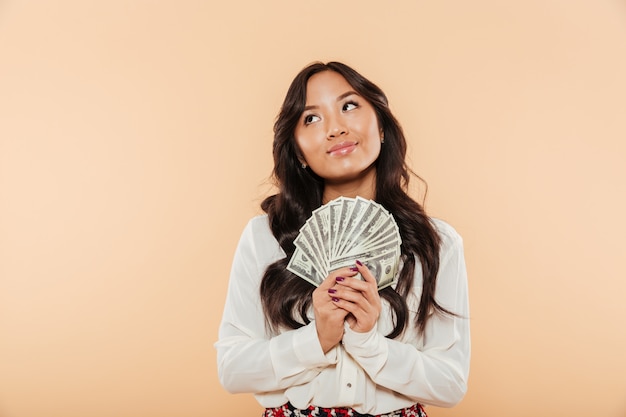 Foto gratuita retrato de mujer asiática morena mirando hacia arriba mientras sostiene el abanico de billetes de 100 dólares siendo exitosa mujer de negocios sobre fondo de durazno