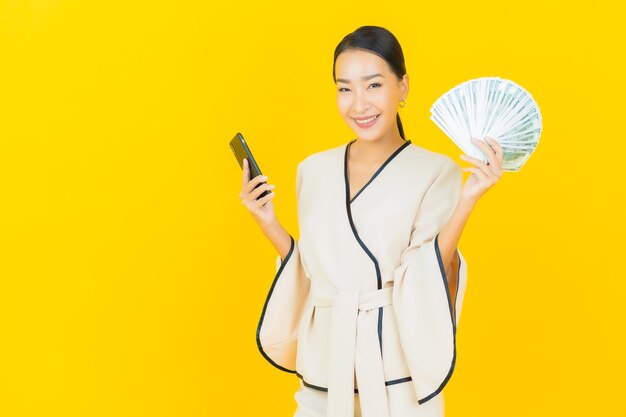 Retrato de mujer asiática de negocios joven hermosa con una gran cantidad de dinero en efectivo y hucha en la pared amarilla