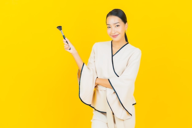 Retrato de mujer asiática de negocios joven hermosa con maquillaje cepillo cosmético en la pared amarilla