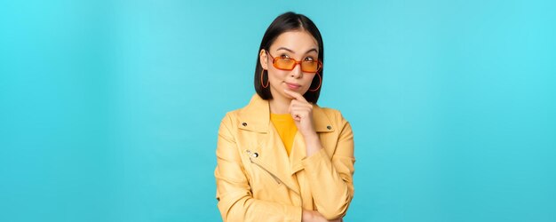 Retrato de una mujer asiática pensando en buscar ideas o soluciones reflexivas con gafas de sol de pie sobre fondo azul.