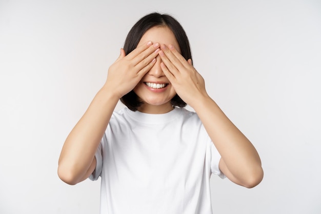 Retrato de mujer asiática que cubre los ojos esperando sorpresa con los ojos vendados sonriendo feliz anticipando estar de pie contra el fondo blanco.