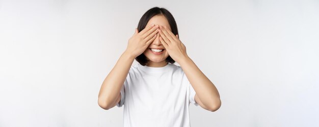 Retrato de mujer asiática que cubre los ojos esperando sorpresa con los ojos vendados sonriendo feliz anticipando estar de pie contra el fondo blanco.