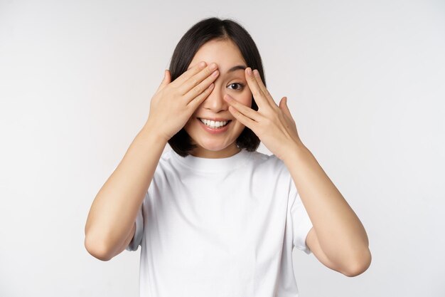 Retrato de mujer asiática que cubre los ojos esperando sorpresa con los ojos vendados sonriendo y mirando a la cámara de pie sobre fondo blanco.