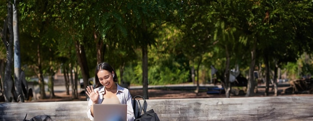 Retrato de una mujer asiática sentada con una computadora portátil en un banco en el parque escuchando música con conexión inalámbrica