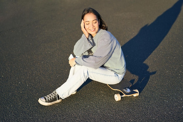 Retrato de mujer asiática sentada en patineta patinando en su longboard de crucero usando la aplicación de teléfono inteligente