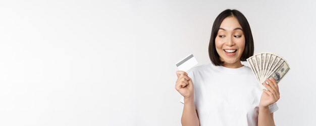 Retrato de mujer asiática sonriendo con tarjeta de crédito y dinero en efectivo dólares de pie en camiseta sobre w...