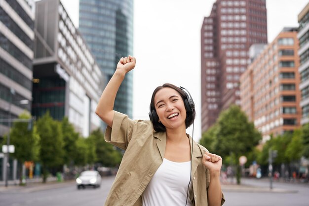 Retrato de mujer asiática sonriente bailando triunfante sintiéndose feliz mientras escucha música en la ciudad posin