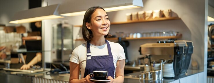 Foto gratuito retrato de una mujer asiática sonriente con delantal barista dándole una taza de café trabajando en una cafetería sirviendo