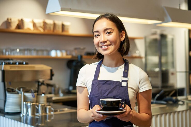 Foto gratuito retrato de una mujer asiática sonriente en delantal barista dándote una taza de café trabajando en un café sirviendo al dr.
