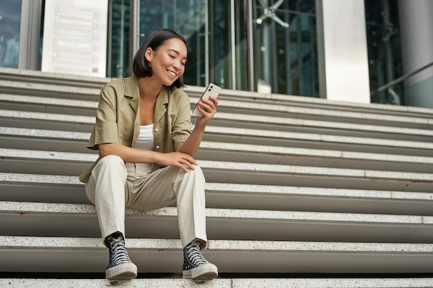 Retrato de una mujer asiática sonriente feliz sentada al aire libre cerca del edificio usando tecnología de teléfono inteligente con