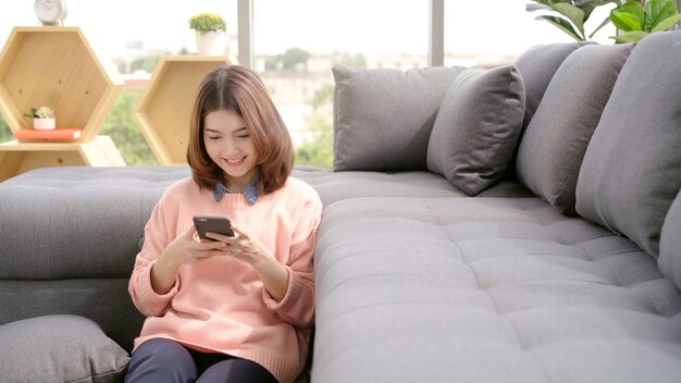 Retrato de la mujer asiática sonriente de los jóvenes atractivos hermosos que usa smartphone mientras que miente en el sofá