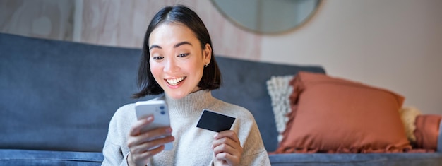 Foto gratuita retrato de una mujer asiática sonriente que paga con tarjeta de crédito en la aplicación de su teléfono inteligente y organiza el débito directo