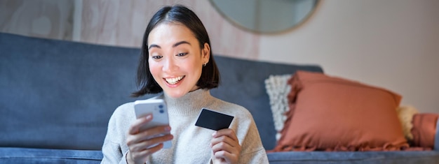 Retrato de una mujer asiática sonriente que paga con tarjeta de crédito en la aplicación de su teléfono inteligente y organiza el débito directo