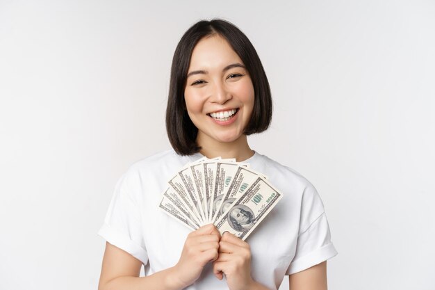 Retrato de una mujer asiática sonriente sosteniendo dólares dinero concepto de financiación de microcréditos y dinero en efectivo de pie sobre fondo blanco.