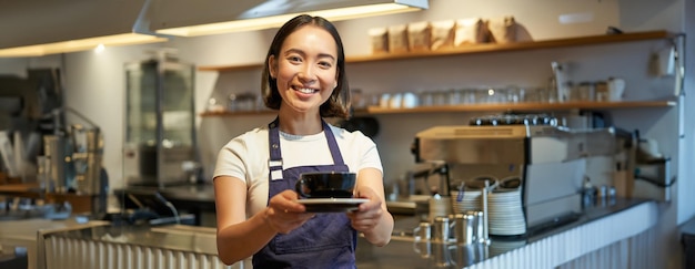 Foto gratuita retrato de una mujer asiática sonriente sostiene una taza de café y prepara bebidas para los clientes en la cafetería trabajando y