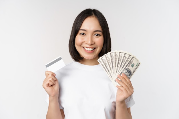 retrato, de, mujer asiática, sonriente, tenencia, tarjeta de crédito, y, dinero efectivo, dólares, posición, en, camiseta, encima, fondo blanco