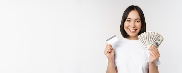 Foto gratuita retrato, de, mujer asiática, sonriente, tenencia, tarjeta de crédito, y, dinero efectivo, dólares, posición, en, camiseta, sobre, w