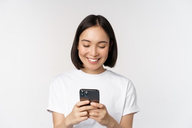 Retrato de una mujer asiática sonriente usando un teléfono móvil chateando mensajes de texto de pie en una camiseta sobre fondo blanco Copiar espacio