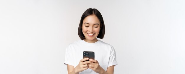 Retrato de una mujer asiática sonriente usando un teléfono móvil chateando mensajes de texto de pie en camiseta sobre fondo blanco