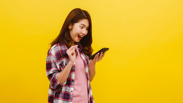 Retrato de mujer asiática sorprendida usando teléfono móvil con expresión positiva, vestido con ropa casual y mirando a cámara sobre pared amarilla. Feliz adorable mujer alegre disfruta el éxito.