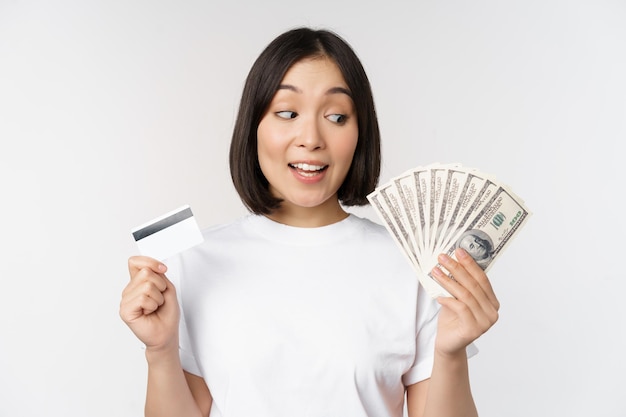 Retrato de mujer asiática sosteniendo dólares de dinero y tarjeta de crédito que parece impresionada y asombrada de pie en camiseta sobre fondo blanco.