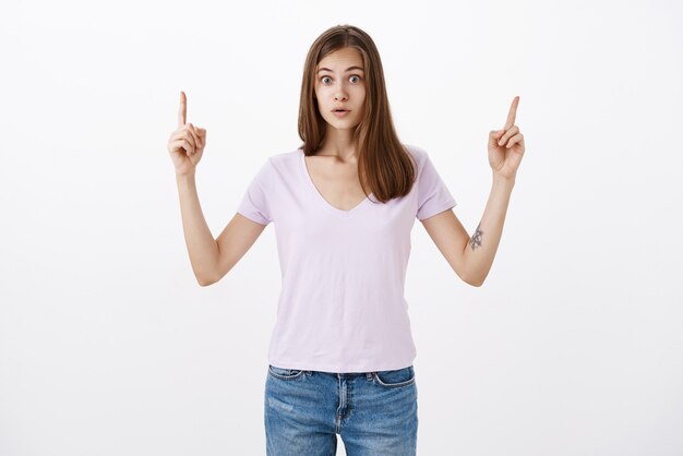 Retrato de mujer asombrada aturdida en camiseta casual con cuello en v levantando los brazos apuntando hacia arriba con asombrada mirada intensa de pie impresionado en estupor