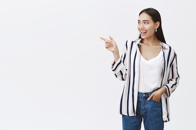 Retrato de una mujer atractiva y amigable con una elegante blusa a rayas y jeans, sosteniendo la mano en el bolsillo, apuntando y mirando hacia la izquierda con una amplia sonrisa