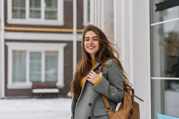 Retrato de mujer atractiva con cabello largo castaño con mochila y una sonrisa suave. Foto de dama caucásica refinada en chaqueta gris posando