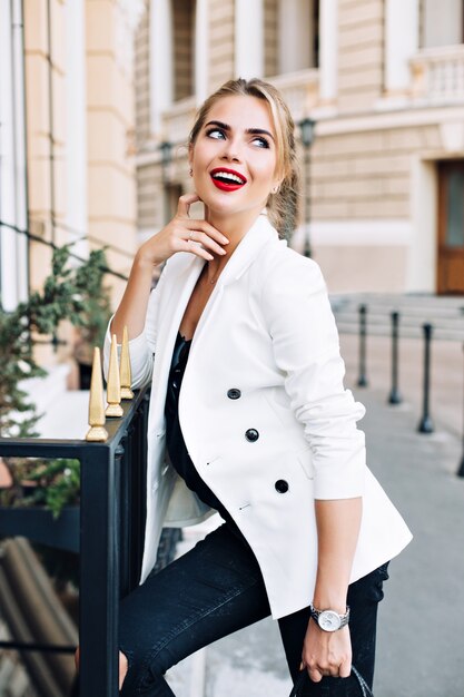 Retrato de mujer atractiva en chaqueta blanca apoyado en la valla en la calle. Ella está sonriendo a un lado.