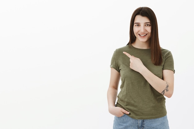 Retrato de mujer atractiva confiada curiosa y optimista con tatuaje sonriendo coqueta y atrevida y apuntando a la esquina superior izquierda mientras pregunta sobre un producto curioso sobre una pared gris