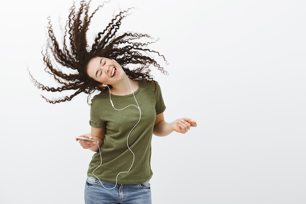 Foto gratuita retrato de mujer atractiva, despreocupada y demasiado emotiva con el pelo rizado, cantando música mientras escucha canciones en auriculares, sosteniendo un teléfono inteligente y disfrutando de grandes cosas