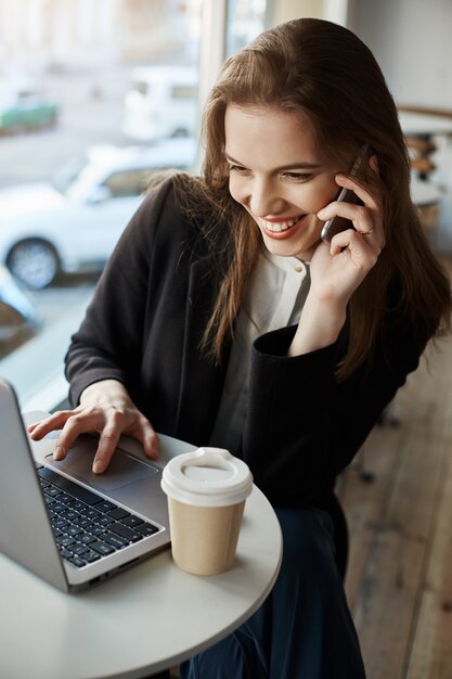 Retrato de mujer atractiva y elegante en la cafetería, navegando en Internet mientras bebe café y habla por teléfono inteligente, trabaja mientras escucha los últimos chismes de un amigo