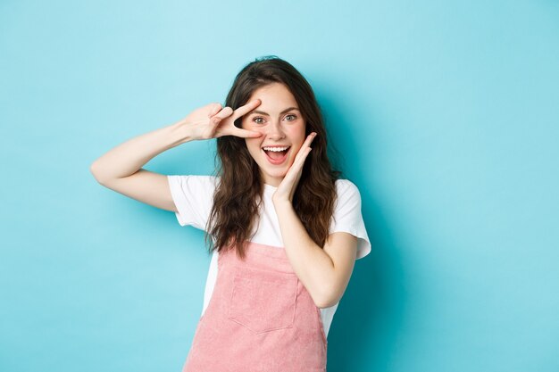 Retrato de mujer atractiva emocionada que muestra el signo v cerca del ojo, haciendo un gesto kawaii y sonriendo feliz a la cámara, de pie optimista contra el fondo azul.