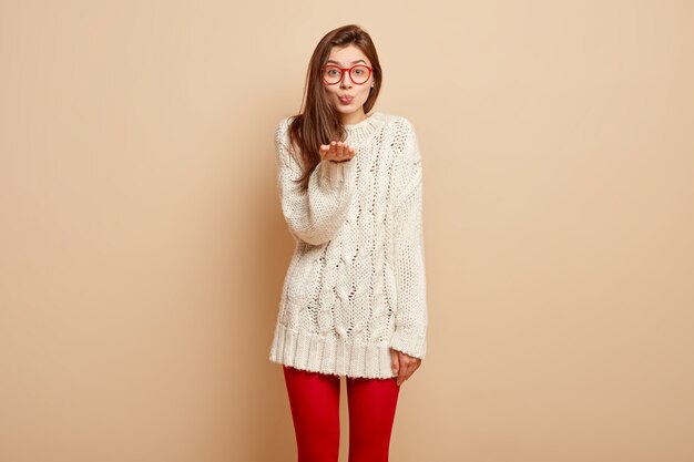 El retrato de una mujer atractiva envía un beso al aire, mantiene las palmas extendidas hacia adelante, viste un jersey de invierno blanco, mallas rojas, expresa amor, tiene los labios fruncidos, demuestra un gesto de saludo o despedida