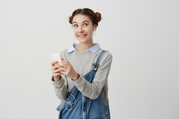 Retrato de mujer atractiva infantil en traje de mezclilla mirando hacia los lados con emociones alegres. Mujer enamorada recibiendo un mensaje agradable en su teléfono inteligente sintiendo felicidad. Expresiones faciales