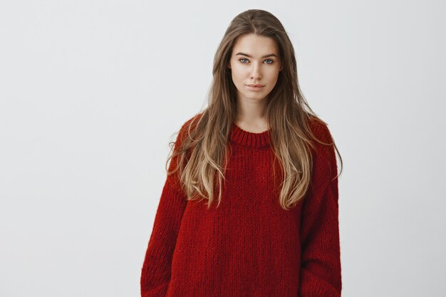 Retrato de mujer atractiva joven sensual de 25 años en suéter rojo acogedor sentado en casa cómodo, disfrutando de mirar la nieve cayendo por las calles, mirando posando contra