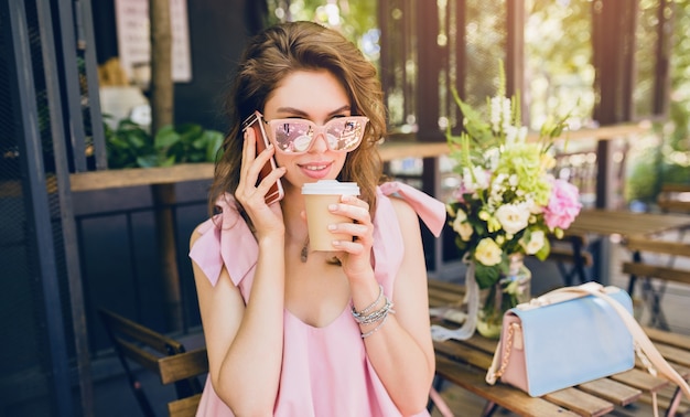 Retrato de mujer atractiva joven sentada en la cafetería, traje de moda de verano, estilo hipster, vestido de algodón rosa, gafas de sol, sonriendo, tomando café, accesorios elegantes, ropa de moda, hablando por teléfono