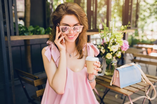 Retrato de mujer atractiva joven sentada en la cafetería, traje de moda de verano, estilo hipster, vestido de algodón rosa, gafas de sol, sonriendo, tomando café, accesorios elegantes, ropa de moda, hablando por teléfono