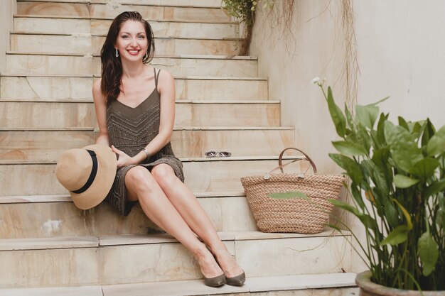 Retrato de mujer atractiva joven sentada en la escalera en elegante vestido con sombrero de paja, estilo veraniego, tendencia de moda, vacaciones, sonriendo, piernas delgadas, accesorios elegantes, bolso