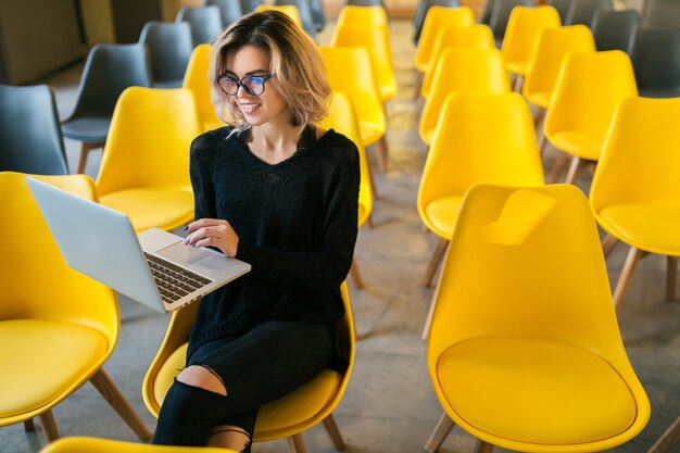 Retrato de mujer atractiva joven sentada en la sala de conferencias trabajando en un portátil con gafas, aprendizaje de los estudiantes en el aula con muchas sillas amarillas