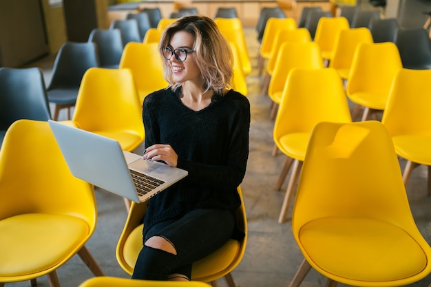 Retrato de mujer atractiva joven sentada en la sala de conferencias trabajando en un portátil con gafas, aprendizaje de los estudiantes en el aula con muchas sillas amarillas