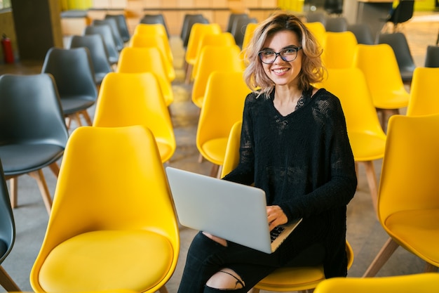 Retrato de mujer atractiva joven sentada en la sala de conferencias trabajando en un portátil con gafas, aprendizaje de los estudiantes en el aula con muchas sillas amarillas