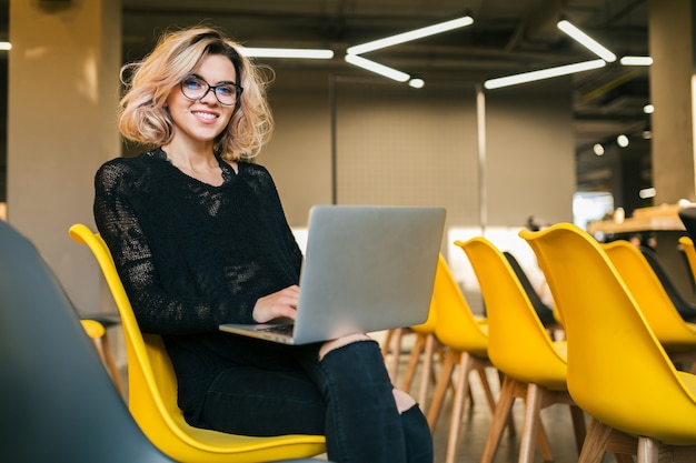 Retrato de mujer atractiva joven sentada en la sala de conferencias trabajando en un portátil con gafas, aprendizaje de los estudiantes en el aula con muchas sillas amarillas