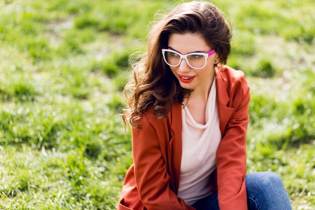 Retrato de mujer atractiva con labios carnosos, anteojos, chaqueta roja, peinado ondulado sentado sobre la hierba verde en el parque soleado de primavera y sonriendo