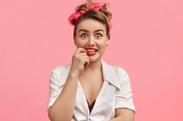 Retrato de mujer atractiva con maquillaje, mantiene el dedo en la boca, tiene dientes blancos y uniformes, usa diadema, blusa blanca, aislado sobre rosa