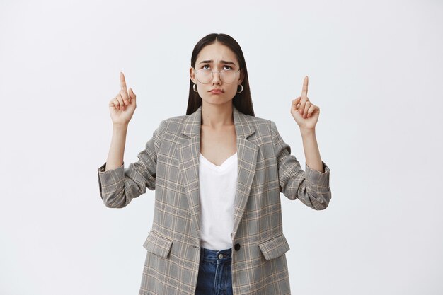 Retrato de mujer atractiva molesta miserable con cabello oscuro y gafas con chaqueta elegante, enfurruñado y lloriqueando mientras apunta y mira hacia arriba, triste y disgustado