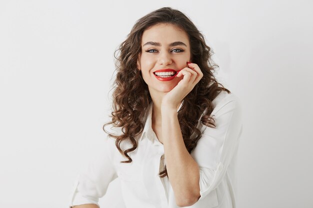 Retrato de mujer atractiva sonriente sincera con dientes blancos aislados, pelo largo y rizado, blusa blanca, estilo de negocios elegante, emoción positiva feliz, maquillaje de lápiz labial rojo de cerca