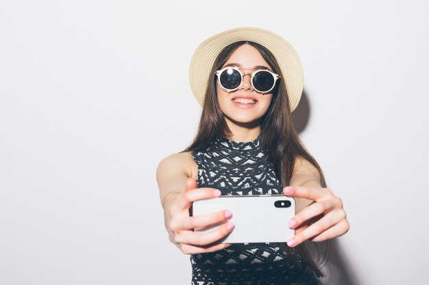 Retrato de mujer atractiva sonriente con sombrero de pie y tomando un selfie aislado